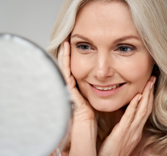 Woman looking into a mirror, smiling with her hands brushing against her cheeks.